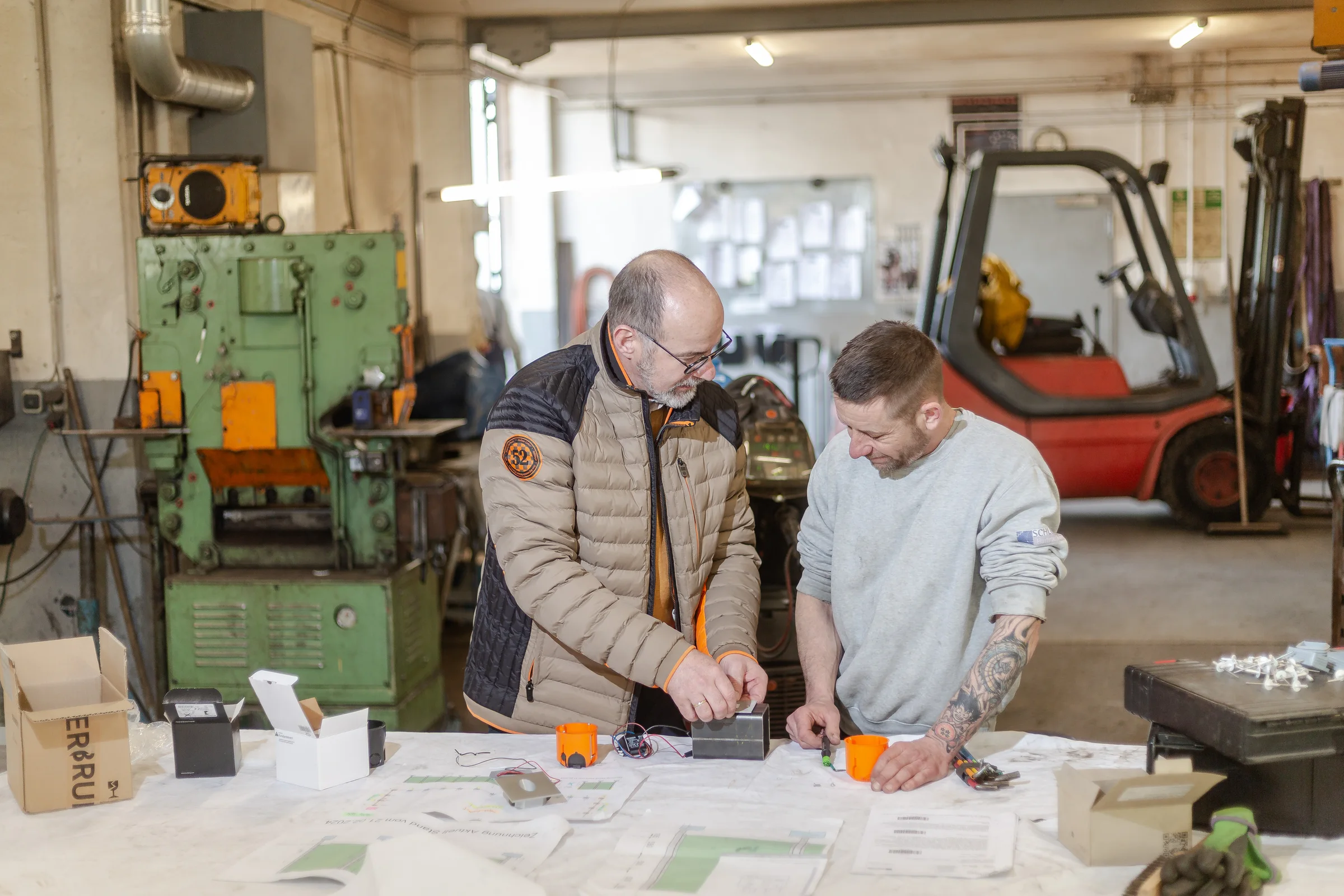 Zwei Mitarbeiter besprechen Bauteile und Unterlagen an einem Arbeitstisch in einer Metallwerkstatt.